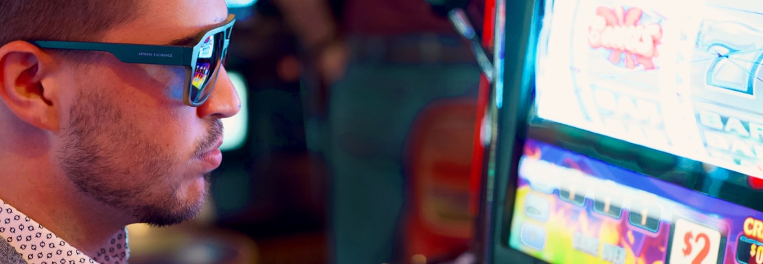 A man wearing sunglasses is intently focused on a slot machine, with colorful lights and graphics illuminating the screen. The scene is set indoors, likely in a casino or gaming area. His attire includes a patterned shirt and a blazer.