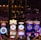 A lively casino scene featuring a row of bright, colorful slot machines with themes like Monopoly, Michael Jackson, and Lord of the Rings. The gaming machines display a variety of flashing lights and digital screens, creating an energetic and exciting atmosphere. Reflections of the lights are visible in glossy surfaces above the machines.